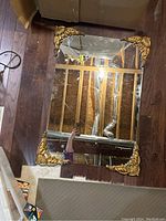 Full view of rectangular clear glass mirror with ornate gold corner embellishments on wooden floor.