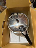 Pressure cooker lid with safety valves and black handle, placed inside original box with foam padding.