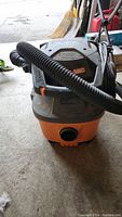 Front view of the Rigid blower vacuum with flexible hose coiled on top, showing orange base and black top with branding.