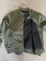 Front view of camo green jacket open to show quilted black interior lining and tag