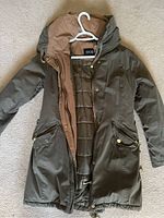 Full view of olive green knee-length jacket with its front open, showing padded inner lining in light brown, zippered pockets with gold buttons, and zipper with gold teeth.