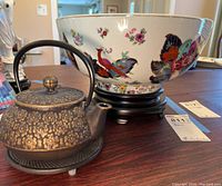 Photo showing metal Japanese teapot on trivet next to decorative floral porcelain bowl on wooden stand.