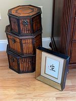 Three stacked octagonal bamboo boxes with black metal latches next to a framed Asian art piece with calligraphy and gold border.