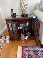 Side table cabinet with glass top, opened double doors show two shelves inside with glass and wooden shelves. Beveled glass doors with brass knobs. Hardwood floor and nearby sofa visible.