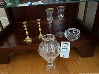 Six candle holders displayed on wooden shelving: two pairs of brass candle holders with octagonal bases; two tall glass candle holders with brass rims; one glass bowl candle holder; one large ornate glass candle holder with bulbous pattern.