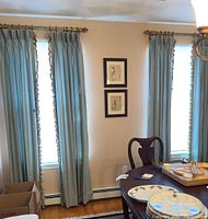 Curtains hanging in front of two windows in a room with a chair and table visible