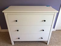 Front view showing three drawers with black knob pulls on a white dresser against a purple wall and carpeted floor.