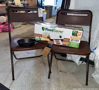 Two brown Cosco folding chairs with metal legs supporting a boxed FoodSaver Vacuum Sealing System and a cast iron skillet.
