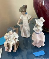 Three porcelain figurines representing girls with dolls and puppies on a blue surface in front of a red vase.