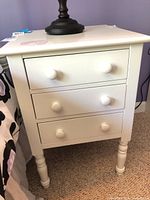 Front view of white nightstand with three drawers, turned legs, placed next to bed.