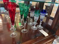 Ten colored glass stemware in red, green, and clear with blue and purple coloring. Displayed on a wooden surface with reflective background.