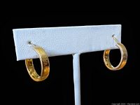 Pair of gold hoops displayed on white earring stand, showing front etched detail