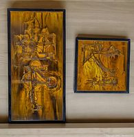 Two wood-framed wall panels, one rectangular and one square, showing abstract brutalist acid-etched and oxidized brass surfaces with geometric and organic shapes in warm metallic tones, against a light wood background.