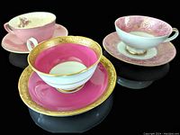 Front view of three vintage pink and gold china teacups with matching saucers, showing the ornate gold detailing and pink tones.