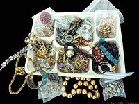 Overhead view of a white tray containing assorted vintage costume jewelry including bracelets, brooches, earrings, bead strands in plastic bags, and mixed metal pieces.