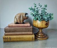 Wood pig figurine on stack of four vintage books alongside a lathe-turned wooden pedestal vase with faux eucalyptus branch inside.