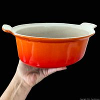 Side view held in hand showing flame orange gradient enameled cast iron casserole dish with cream interior and handles.