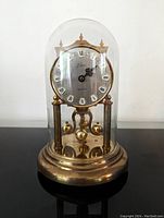 Front view of the upright vintage Kern clock under clear glass dome on brass base with rotating pendulum.