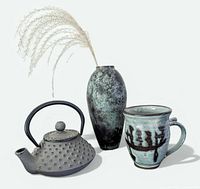 Front angle view of the small cast iron teapot, pottery mug, patinated metal vase with pampas grass stem on white background.