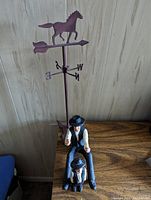 Amish-themed figurine of man and child with attached metal weather vane featuring a horse silhouette and directional letters.