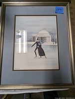 Photo of framed artwork showing ice skating couple in front of classical domed building in winter scene, blue and gray tones, framed in silver metal with mats.