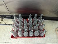 Top-down view of 23 empty 'Drink Me Pop' glass bottles arranged in red plastic carrier crate.