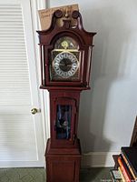 Front view of the tall wooden grandmother clock showing the clock face with Roman numerals and decorative brass details, the arched top with carved scrolls, and lower glass panel with pendulum.
