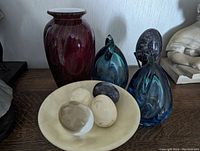 Photo showing burgundy glass vase, two blue glass bird figurines, ceramic bowl with three eggs of glass and ceramic.