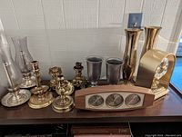 Photo showing brass and pewter candlestick holders, pewter cups, brass vases, brass clock, and wood base weather station with clock, thermometer, and barometer.