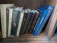 Books sitting upright on a wooden shelf, titles visible on spines showing a collection of large, hardcover coffee table books.