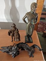 Three statuettes displayed together on a wooden surface: tall bronze figure, couple on horse, dark dragon figure