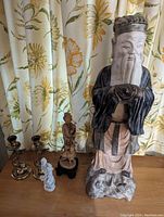 Full view of the tall Oriental statue flanked by other smaller statuettes and brass candle sticks on a wooden surface with floral curtains in background.