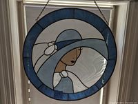 Close-up photo showing the stained glass artwork of a woman in a blue hat with a white flower and detailed facial features, framed with blue and clear glass sections.