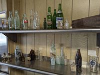 Two metal shelves with a wide variety of vintage bottles of different sizes, glass beer bottles, and a wooden cheese box on the top shelf.