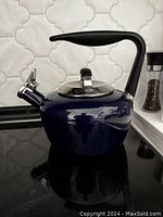 Glossy blue enamel kettle with a curved black plastic handle and a silver metal lid with a black knob. The kettle is on a black stovetop, showing the spout with a silver cover.