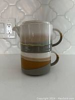 Stacked view of ceramic tea pot and mug showing side handle and spout with multicolor bands in grey, white, and yellow.