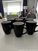 Front view showing eight black coffee mugs on a white surface with light coming from window background