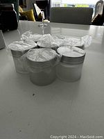 View of six brand new frosted glass cream jars with silver lids wrapped in plastic protection, arranged together on a white surface.
