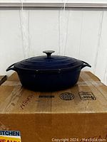 Front view of oval blue cast iron Dutch oven with lid on a cardboard box showing signs of wear.