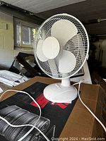 White tabletop electric fan positioned on a cardboard box in a storage area, showing front view with control buttons and fan blades visible.