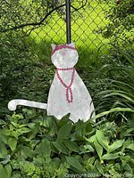 Wooden cat-shaped sign standing in outdoor garden area among greenery. Painted white with pink necklace and bow details.