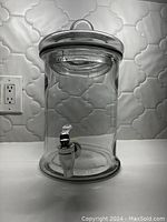 Front view of the clear glass water dispenser showing the metal lid with loop handle and chrome spigot near base.