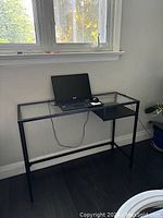 Full view of black metal frame desk with glass surface near window and a laptop placed on top.