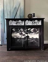 Front view of black wooden cupboard showing white decorative transfers on two drawers and scenic landscape transfers on two cabinet doors.