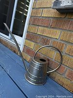 Metal watering can placed on a blue wooden surface next to a brick wall.