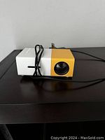 Photo of the outdoor projector, showing its white and yellow casing with a front lens and attached power cable.