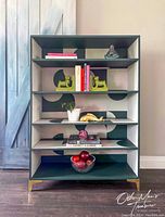 Front view of the bookcase showing abstract geometric painted design in green and pink with some black and white. The shelves hold decorative items which are not included.