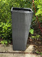 Photo showing full view of tall rectangular grey planter with wicker texture outdoors on a stone surface next to chain link fence and greenery.