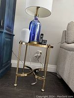 Round brass and glass side table shown next to couch and cabinet, holding a lamp and other electronic items (not included).