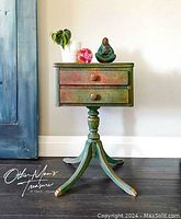 Full view of small side table showing two drawers, turned pedestal leg and tripod base. Painted in blended tones of green, gold and red.
